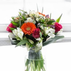 A testament to local artistry, this exquisite floral arrangement radiates from a sunlit corner of Walworth, alive with the harmonious play of color, structure, and emotion. Creamy peach gerberas form a luminous halo around the bouquet's core, their petals unfolding with gentle grace, while vivid orange gerbera blossoms infuse energy, reminiscent of festive lanterns lighting up Westmoreland Road. Intertwined are bold fuchsia roses, their saturated hues adding romance and sophistication, contrasted beautifully with clusters of crisp white alstroemeria and small spray roses-which shimmer like morning dew atop Camberwell Green. The arrangement's depth is heightened by hints of light pink blooms and fine, delicate white fillers that evoke a sense of ongoing abundance. The foundation is a cascade of glossy, broad aspidistra leaves-curved and overlapping protectively around the bouquet's base-paired with slender, pointed branches of fresh greenery. Together, they give structure and lush tranquility, grounding the dazzling florals above. All are gathered in a clear glass vase, where each green stem is visible, reinforcing the design's natural freshness. The blooms seem to glow against a luminous, minimalist backdrop, capturing the serenity and happiness of a new beginning in Walworth. The mixed scent-faintly sweet from the roses and lilies, fresh from the foliage-lends a tranquil, uplifting air, making this piece a perfect centrepiece for gatherings, celebrations, or welcoming someone home to the heart of Walworth.